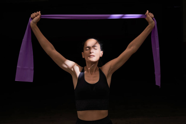 Apto atleta feminina com raios de sol no rosto em sportswear de pé com os braços estendidos durante o exercício com banda de resistência roxa durante o treinamento em sala escura - Foto, Imagem