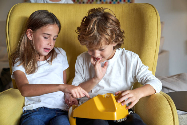 Nette Kinder in Freizeitkleidung wählen die Nummer auf einem gelben Telefon, während sie zusammen in einem bequemen Sessel in einem hellen Raum sitzen - Foto, Bild