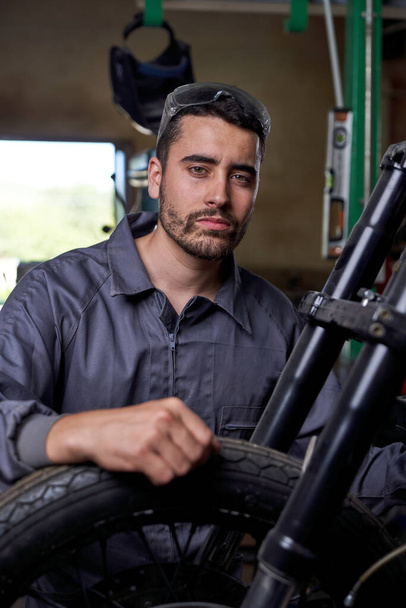 Confident bearded man in uniform sitting near motorcycle with hand on wheel and looking at camera while working in professional workshop - Photo, Image