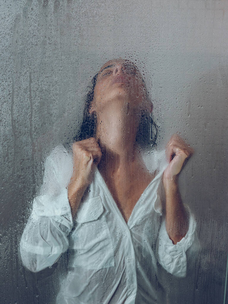 Durch das Glas einer attraktiven Frau in nasser Bluse, die in einer hellen Duschkabine mit transparenter Tür und Wassertropfen im Badezimmer steht - Foto, Bild