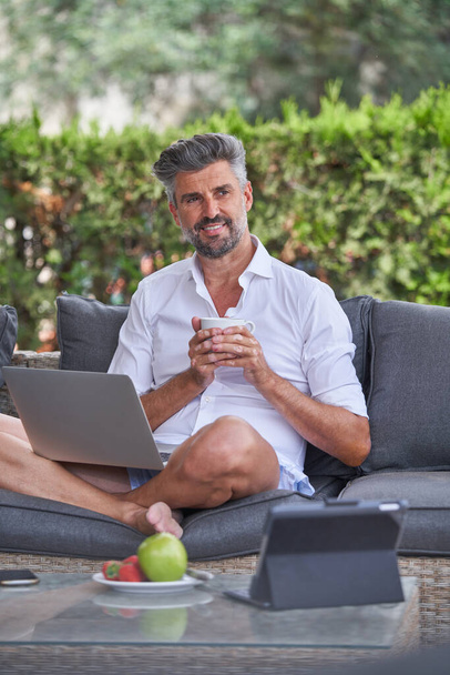 Parrakas mies freelancer harmaat hiukset hymyilee ja katselee pois juodessaan kahvia ja työskentelee etänä kannettavan tietokoneen pihalla - Valokuva, kuva