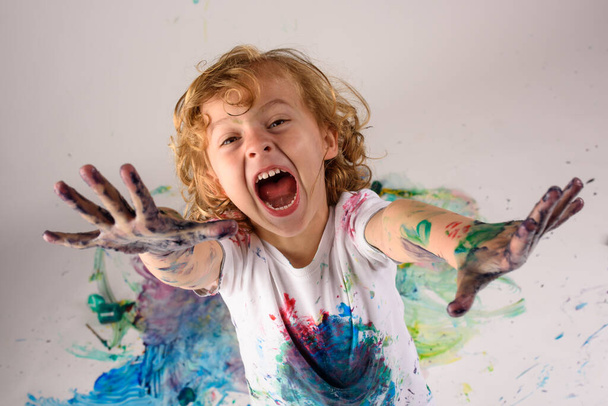 D'en haut du garçon drôle recouvert de peintures colorées regardant la caméra avec le visage excité sur fond blanc dans un studio désordonné - Photo, image