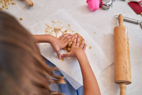 自宅の軽いキッチンで木製のローリングピンでテーブルにクッキー生地を準備する匿名の子供の上から - 写真・画像