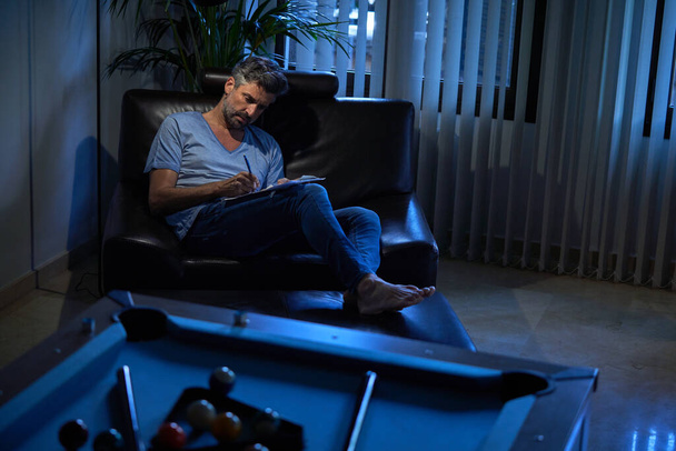 Plná délka podnikatele v ležérní oblečení psaní poznámek při odpočinku v pohodlném křesle v útulné kanceláři v noci - Fotografie, Obrázek