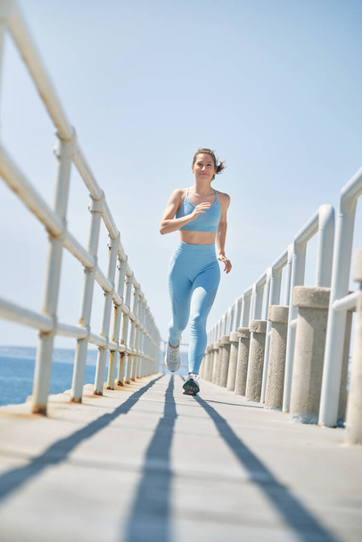 Veselý ženský atlet ve sportovním oblečení jogging na oploceném mostě s odstíny při pohledu na kameru při cvičení - Fotografie, Obrázek