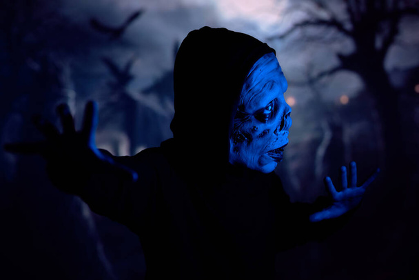 Griezelig monster gesticuleren en voorzichtig weg te kijken terwijl sluipen door donkere begraafplaats onder blauw neon licht - Foto, afbeelding
