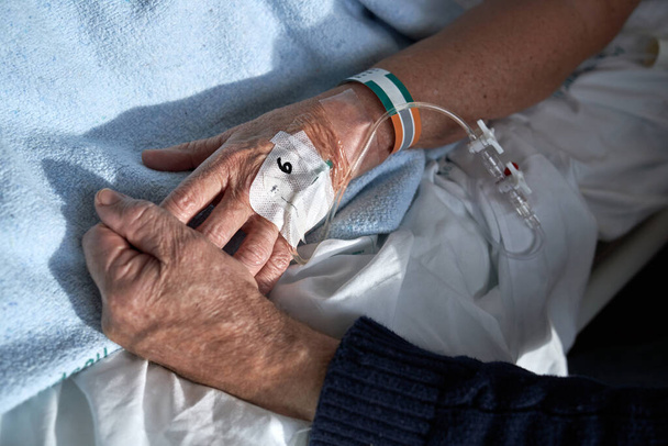 Von oben nicht wiederzuerkennender älterer Mann, der die Hand seiner kranken Frau hält, die während der Behandlung im Krankenhaus auf dem Krankenbett mit angeschlossenem peripheren Venenkatheter liegt - Foto, Bild