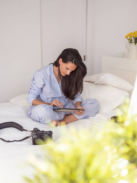 Full body of barefoot focused brunette in pajamas sitting on bed legs crossed near photo camera and using tablet while resting at home - Photo, Image