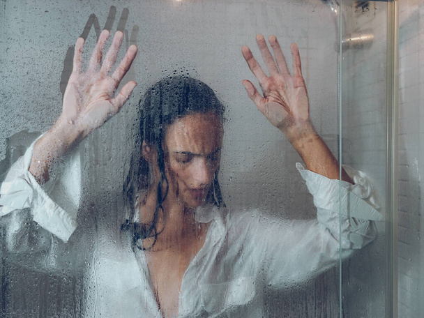 À travers le verre de femme triste avec les cheveux noirs dans l'usure humide touchant porte transparente de cabine de douche dans la salle de bain légère - Photo, image