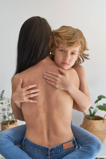 Peaceful kid curly hair and with closed eyes tenderly embracing anonymous topless mother while sitting in light room at home  - Photo, Image