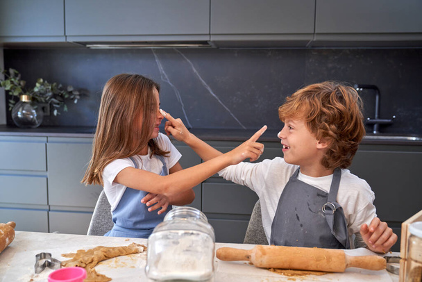 Frère et sœur joyeux se touchant le nez avec des index sales de farine tout en pétrissant la pâte assis au comptoir de la cuisine - Photo, image