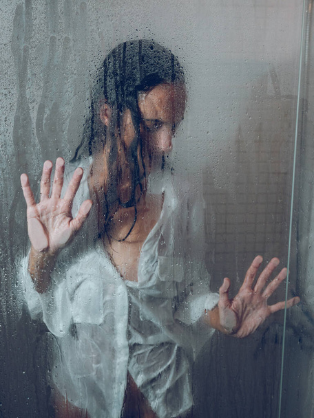 毎日のルーチンの間にシャワーキャビンに立っている間透明なぬれたドアに触れるぬれた毛が付いている敏感な女性のガラスを通して - 写真・画像