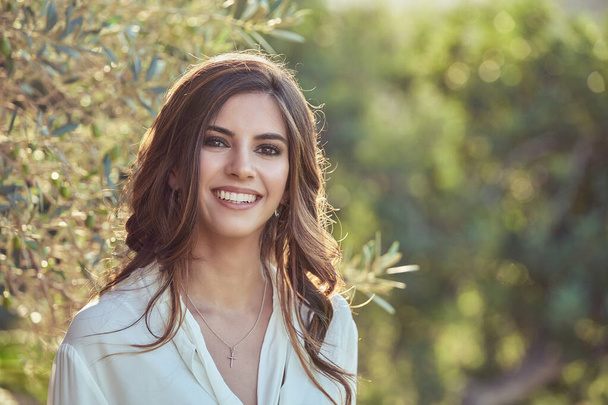 Jeune femme optimiste aux cheveux bouclés regardant la caméra avec le sourire tout en se tenant près d'un arbre vert dans la campagne sur fond flou - Photo, image