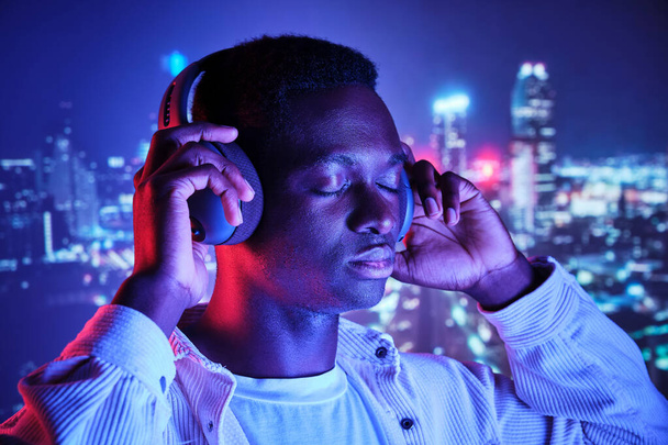 Spokojny Afroamerykanin z zamkniętymi oczami słuchający piosenek w bezprzewodowych słuchawkach przeciwko projekcji świateł miasta - Zdjęcie, obraz