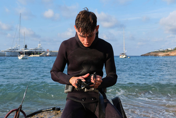 Mergulhador masculino sério em roupa de mergulho sentado em pedra perto do arpão olhando para baixo enquanto limpa óculos de mergulho antes de mergulhar no mar azul - Foto, Imagem