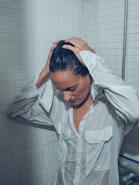 Fatigue femelle en chemise mouillée debout sous les éclaboussures d'eau dans la cabine de douche et toucher la tête - Photo, image