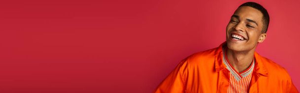 excited african american guy laughing on red background, orange shirt, portrait, banner, copy space - Foto, Bild