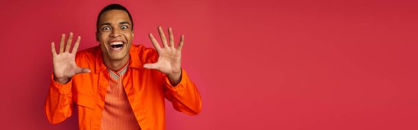 смішний афроамериканський хлопець, що показує страшний жест і посміхається на червоній, помаранчевій сорочці, банер - Фото, зображення
