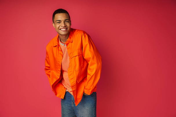 trendy african american man with hands in pocket smiling at camera on red background - Foto, Imagen
