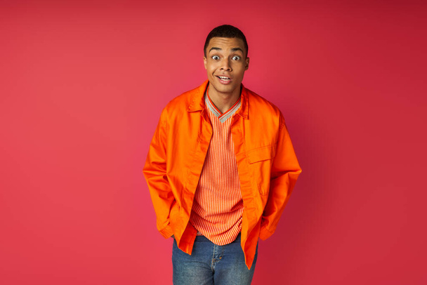 surprised african american man with hands in pocket, in orange shirt looking at camera on red - Fotó, kép