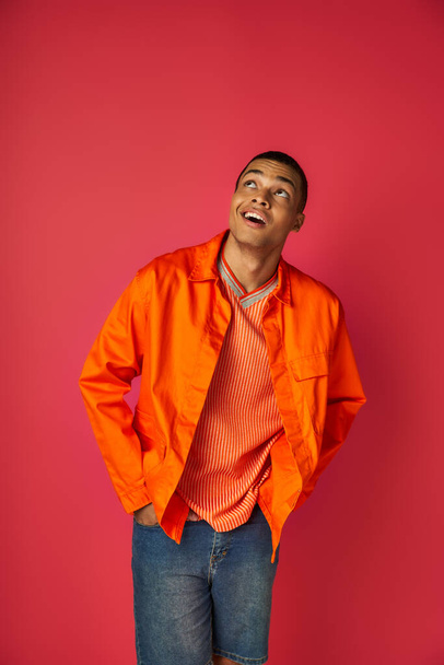 positive and curious african american in orange shirt looking up on red background, hands in pockets - Fotografie, Obrázek