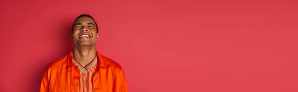 chico afroamericano emocionado riendo con los ojos cerrados en rojo, camisa naranja, pancarta, espacio de copia - Foto, Imagen