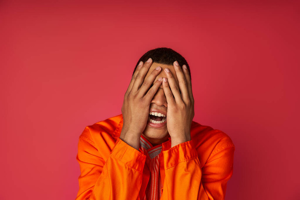 νεαρός καταθλιπτικός Αφροαμερικανός με πορτοκαλί πουκάμισο σκιάζει το πρόσωπο με τα χέρια στο κόκκινο φόντο - Φωτογραφία, εικόνα