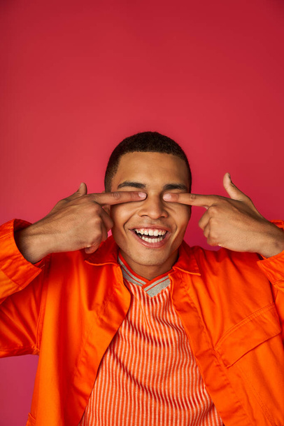 cheerful african american guy obscuring eyes with fingers, orange shirt, red background - Foto, immagini
