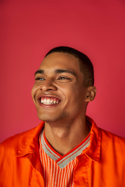 portrait of youthful african american man with radiant smile, stylish orange shirt, red background - Photo, Image