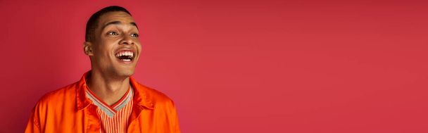 astonished african american man, laughing, looking away, orange shirt, red background, banner - Foto, imagen