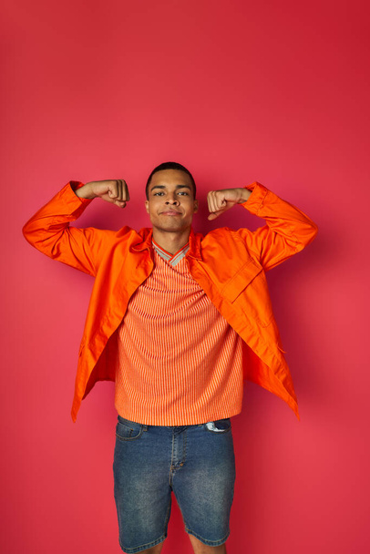 オレンジ色のシャツを着たアフリカ系アメリカ人男性が,赤い背景に強さを示しています. - 写真・画像