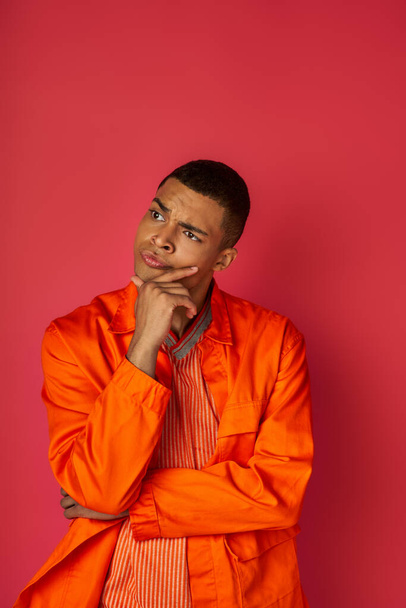 deep in thought african american man in orange shirt touching chest and looking away on red - Фото, зображення