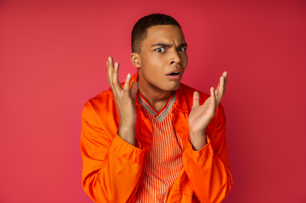 displeased, shocked african american in orange shirt gesturing, looking at camera on red background - Photo, Image