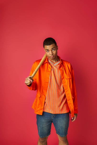 confident african american man in orange shirt holding baseball bat and looking at camera on red - Фото, изображение
