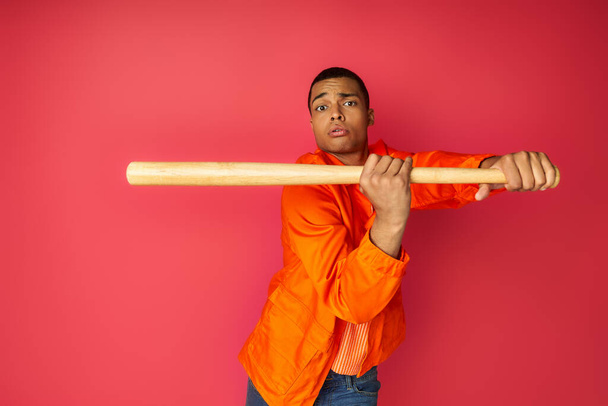 concentrated african american man in orange shirt holding baseball bat and looking at camera on red - Foto, Imagen