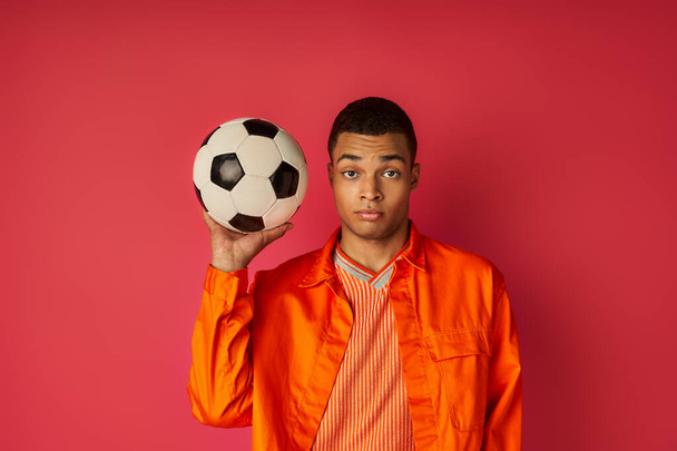 youthful african american guy in orange shirt holding soccer ball and looking at camera on red - Fotó, kép