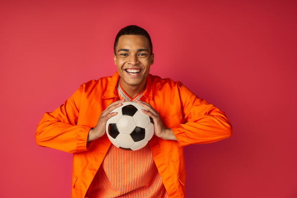 joyful african american man in orange shirt holding soccer ball and smiling at camera on red - Photo, Image