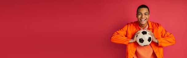 Χαρούμενος Αφροαμερικάνος με πορτοκαλί πουκάμισο κρατώντας μπάλα ποδοσφαίρου σε κόκκινο, λάβαρο, αντίγραφο χώρου - Φωτογραφία, εικόνα