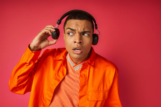 здивований афроамериканський хлопець в помаранчевій сорочці слухає музику в бездротових навушниках на червоному - Фото, зображення