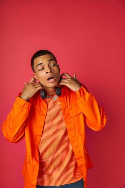 hombre afroamericano sorprendido en camisa naranja, con auriculares inalámbricos mirando hacia otro lado en rojo - Foto, imagen