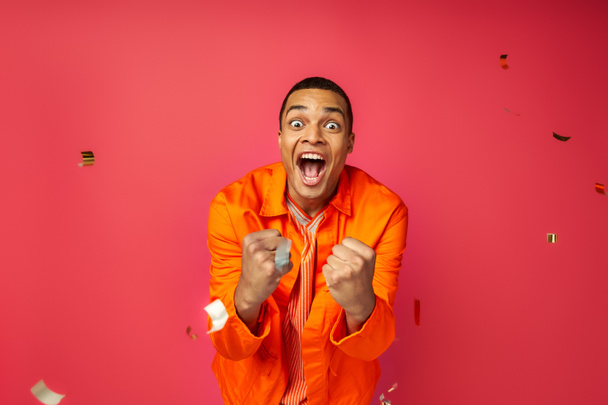 overjoyed african american man in orange shirt shouting and showing win gesture near confetti on red - Foto, Bild