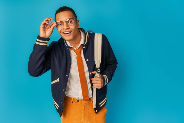 stylish african american student in eyeglasses smiling at camera on blue, notebooks, backpack - 写真・画像
