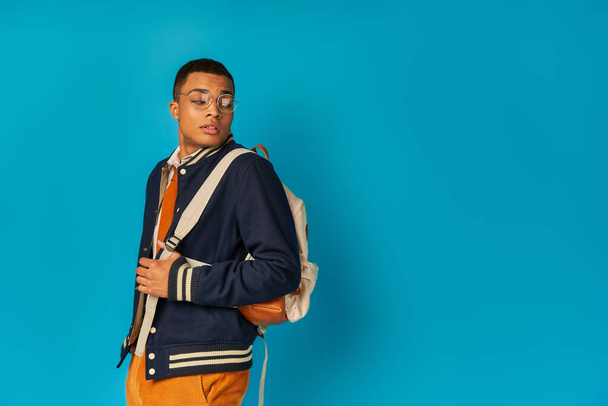 trendy african american student in eyeglasses and jacket looking at backpack on blue background - 写真・画像
