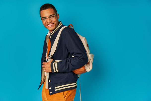 Unbekümmerter afrikanisch-amerikanischer Student in Jacke und orangefarbener Hose, mit Rucksack in die Kamera blickend auf blau - Foto, Bild