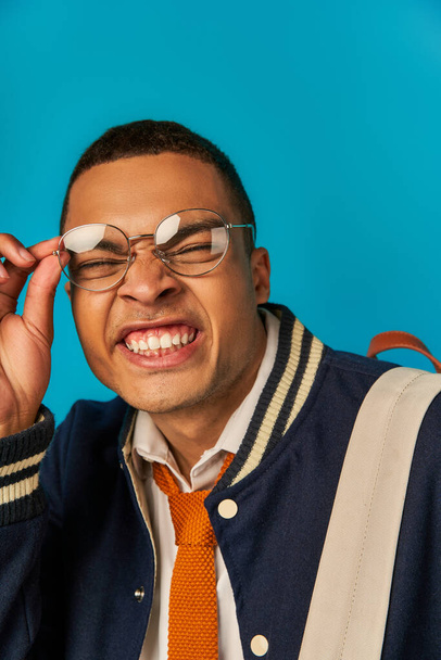 funny african american student in jacket adjusting eyeglasses and grimacing on blue background - Foto, Imagen