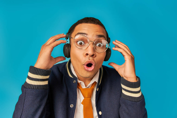 impressed african american student in eyeglasses and headphones, with open mouth on blue - 写真・画像