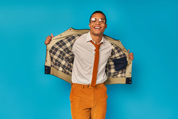 cheerful african american student in trendy clothes opening jacket and smiling at camera on blue - Фото, изображение