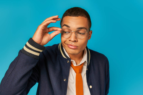 estudiante afroamericano escéptico en chaqueta elegante ajustando gafas y mirando hacia otro lado en azul - Foto, Imagen