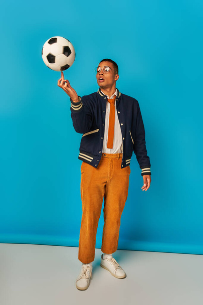 african american student in jacket and orange pants playing with soccer ball on blue - Photo, Image