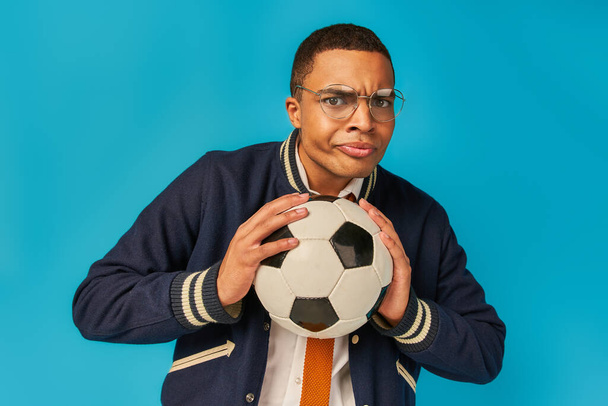 σοβαρός Αφροαμερικάνος φοιτητής με γυαλιά που κρατάει μπάλα ποδοσφαίρου και φουσκώνει μάγουλα στο μπλε - Φωτογραφία, εικόνα